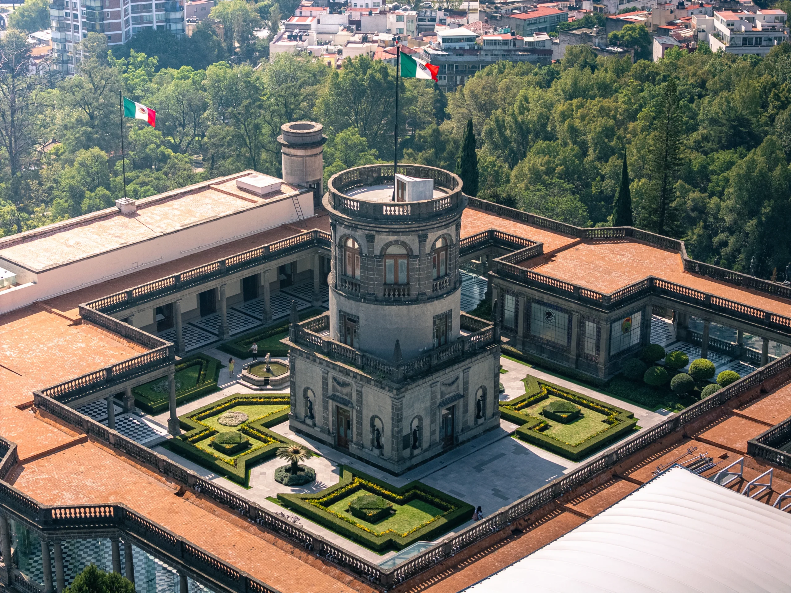 Vista aérea del Castillo de Chapultepec con su torre central, jardines geométricos, banderas mexicanas y edificios históricos rodeados de áreas verdes en la CDMX.