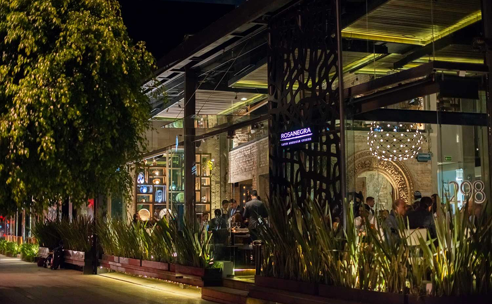 Restaurante moderno y elegante en Polanco de noche, con fachada de cristal iluminada, vegetación decorativa y personas disfrutando una cena en una zona peatonal segura y caminable de la Ciudad de México.