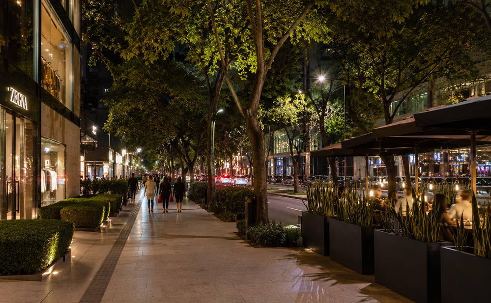 Calle arbolada y amplia banqueta iluminada por la noche en Polanco con personas caminando, escaparates de tiendas y terrazas de restaurantes.
